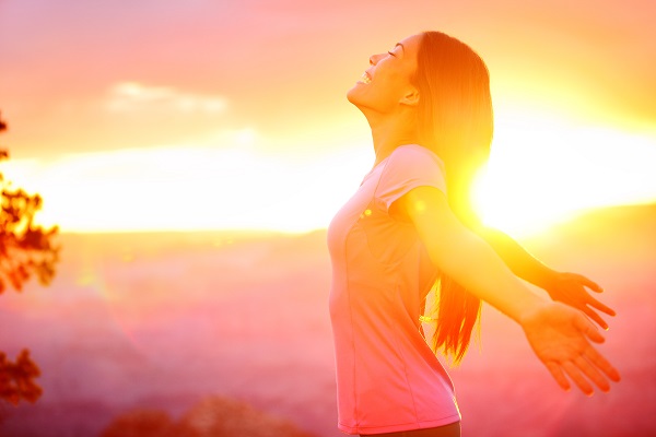 Woman in profile standing in front of a sunrise looking happy and relaxed with outstretched arms